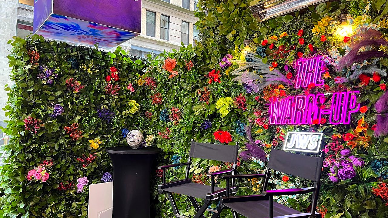 Greenery and flower wall with a pink neon sign and director chairs at a live event