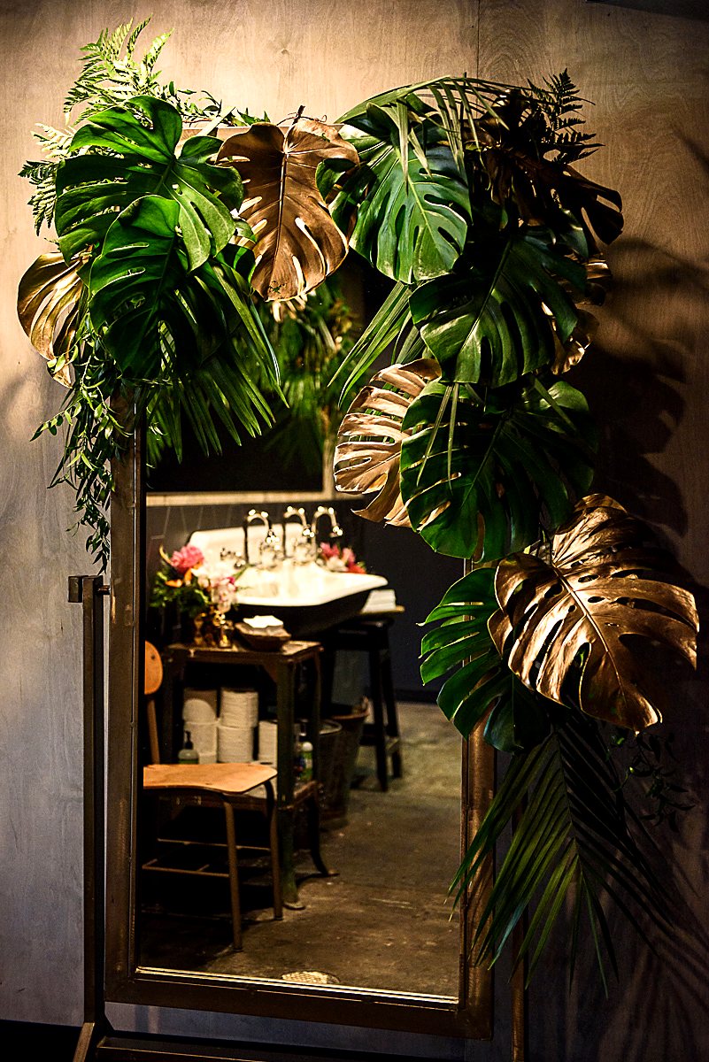 Monstera leaves, some gold-dipped, framing a mirror in a restaurant restroom