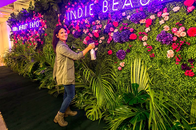Carly Cylinder misting a pink and purple flower wall with neon signs at Indie Beauty Expo