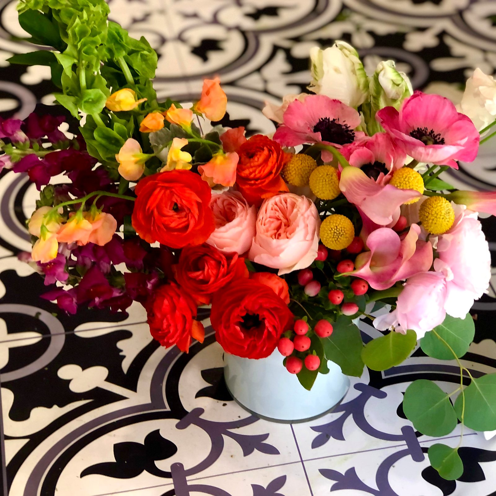 Red and orange ranunculus, pink anemones, craspedia and garden roses on patterned tile