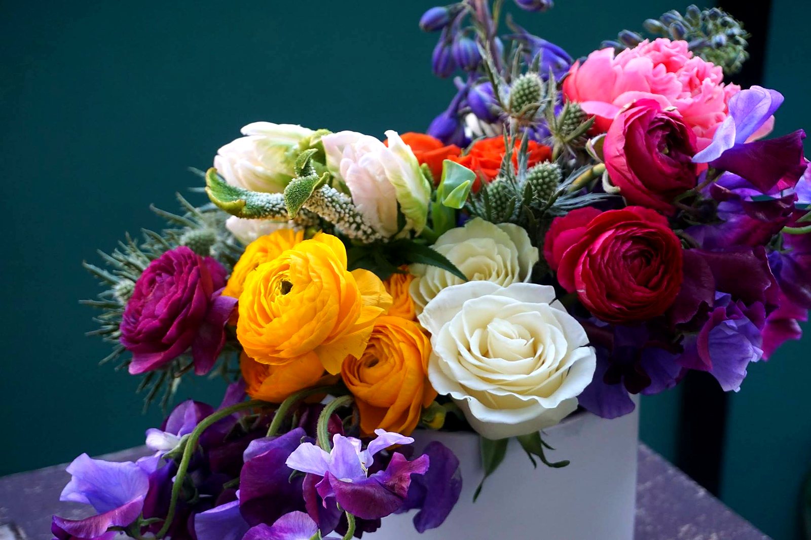Colorful arrangement of ranunculus, white roses, sweet pea and a pink peony against a teal wall