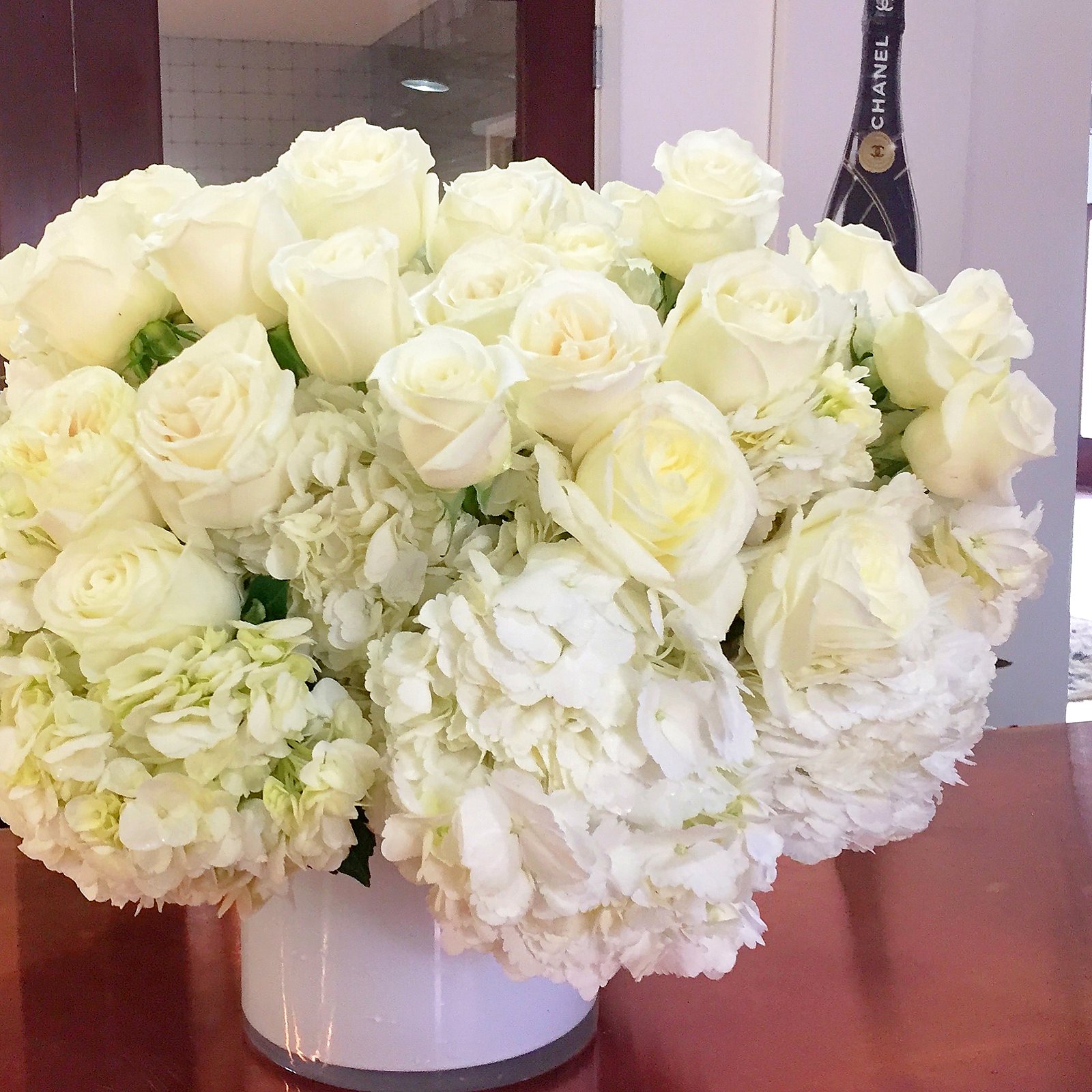 Dense white rose and hydrangea arrangement in a white vase with a Chanel bottle in the background